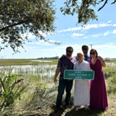 four people stand beside a marsh holding a sign that reads sign reading "George J. 'Geordie' Wallinger, III Bridge"