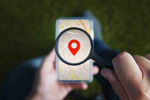 Shot of male hands holding magnifying glass with the red icon of geo-location and looking through it to the smartphone with online city map on the screen